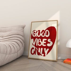 May include: A framed print with a red heart shape containing the text "GOOD VIBES ONLY" in white lettering. The print is hanging on a white wall.