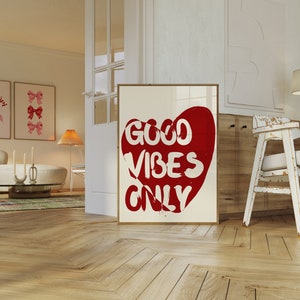 May include: A white framed poster with a red heart shape containing the text "GOOD VIBES ONLY". The poster is hanging on a white wall in a room with wooden floors.