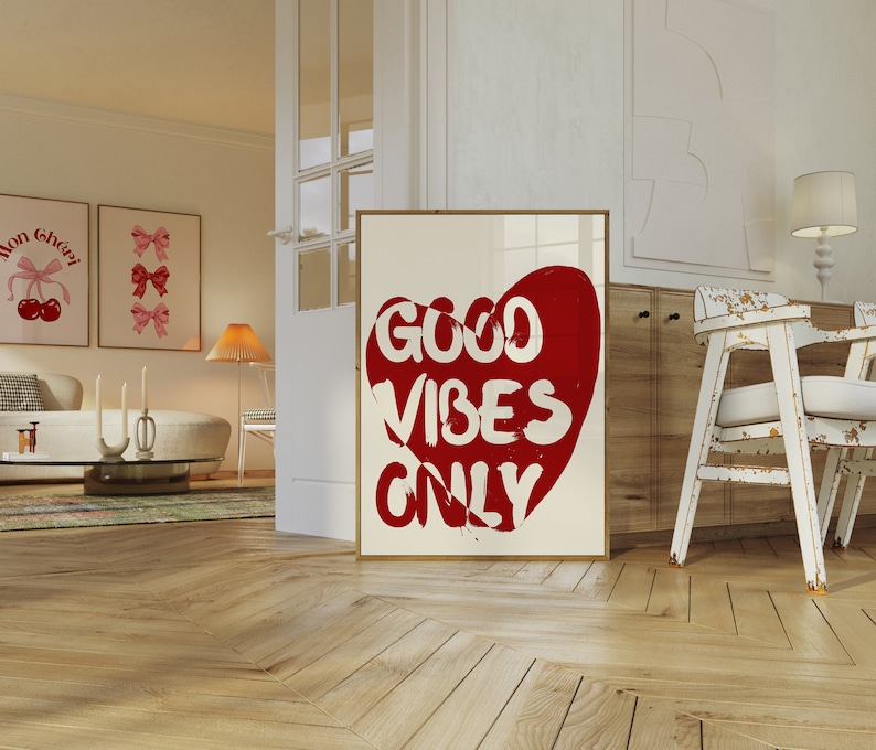 May include: A white framed poster with a red heart shape containing the text "GOOD VIBES ONLY". The poster is hanging on a white wall in a room with wooden floors.
