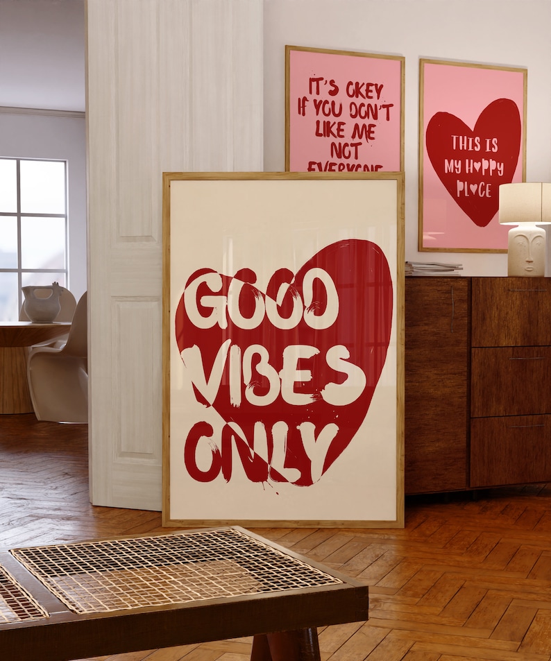 May include: A white framed poster with a red heart shape containing the text "GOOD VIBES ONLY" in white. The poster is hanging on a wall with two other posters, one with the text "IT'S OKEY IF YOU DON'T LIKE ME NOT EVERYONE" and the other with the text "THIS IS MY HAPPY PLACE" with a red heart.