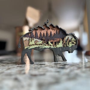 Handcrafted wooden bison wall art featuring a layered forest and mountain scene with wildlife details. Measures 6"x6"x3/8", perfect for rustic, farmhouse, or cabin décor. A unique gift for nature lovers and wildlife enthusiasts.