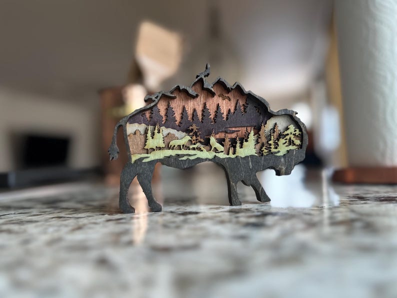 Handcrafted wooden bison wall art featuring a layered forest and mountain scene with wildlife details. Measures 6"x6"x3/8", perfect for rustic, farmhouse, or cabin décor. A unique gift for nature lovers and wildlife enthusiasts.