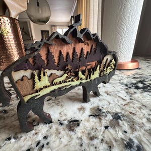 Handcrafted wooden bison wall art featuring a layered forest and mountain scene with wildlife details. Measures 6"x6"x3/8", perfect for rustic, farmhouse, or cabin décor. A unique gift for nature lovers and wildlife enthusiasts.
