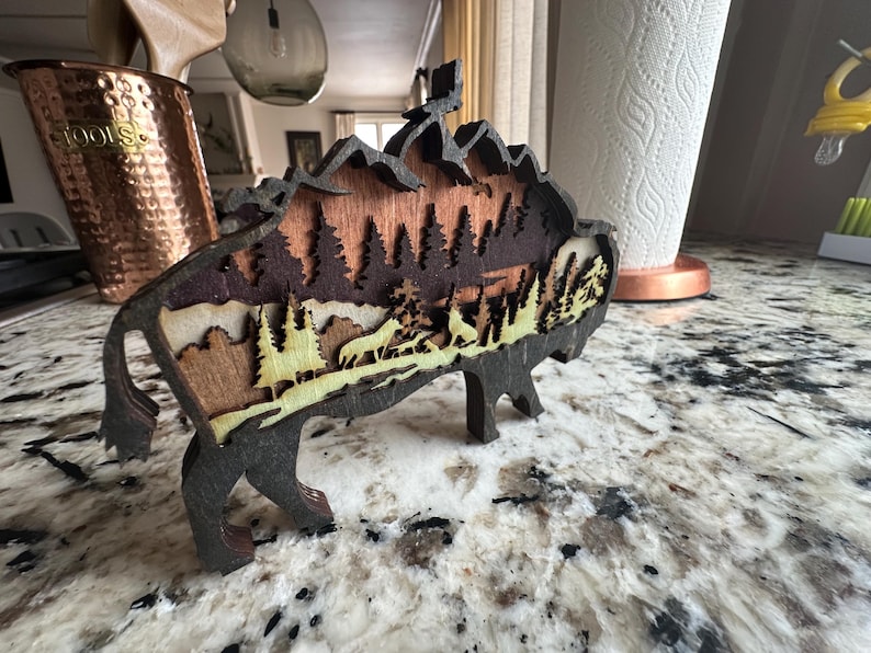 Handcrafted wooden bison wall art featuring a layered forest and mountain scene with wildlife details. Measures 6"x6"x3/8", perfect for rustic, farmhouse, or cabin décor. A unique gift for nature lovers and wildlife enthusiasts.