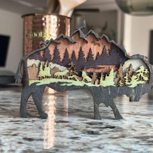 Handcrafted wooden bison wall art featuring a layered forest and mountain scene with wildlife details. Measures 6"x6"x3/8", perfect for rustic, farmhouse, or cabin décor. A unique gift for nature lovers and wildlife enthusiasts.