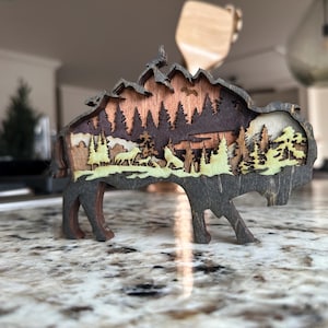 Handcrafted wooden bison wall art featuring a layered forest and mountain scene with wildlife details. Measures 6"x6"x3/8", perfect for rustic, farmhouse, or cabin décor. A unique gift for nature lovers and wildlife enthusiasts.