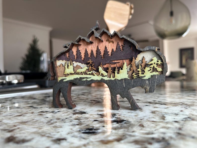 Handcrafted wooden bison wall art featuring a layered forest and mountain scene with wildlife details. Measures 6"x6"x3/8", perfect for rustic, farmhouse, or cabin décor. A unique gift for nature lovers and wildlife enthusiasts.