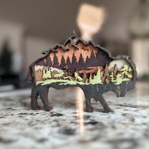 Handcrafted wooden bison wall art featuring a layered forest and mountain scene with wildlife details. Measures 6"x6"x3/8", perfect for rustic, farmhouse, or cabin décor. A unique gift for nature lovers and wildlife enthusiasts.