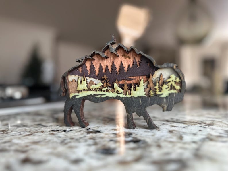 Handcrafted wooden bison wall art featuring a layered forest and mountain scene with wildlife details. Measures 6"x6"x3/8", perfect for rustic, farmhouse, or cabin décor. A unique gift for nature lovers and wildlife enthusiasts.