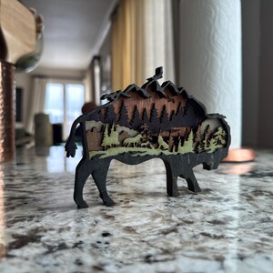 Handcrafted wooden bison wall art featuring a layered forest and mountain scene with wildlife details. Measures 6"x6"x3/8", perfect for rustic, farmhouse, or cabin décor. A unique gift for nature lovers and wildlife enthusiasts.