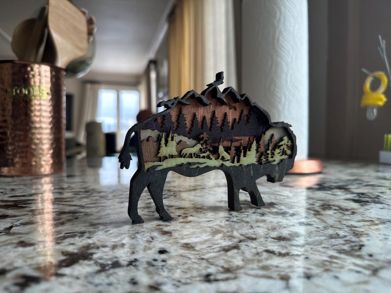 Handcrafted wooden bison wall art featuring a layered forest and mountain scene with wildlife details. Measures 6"x6"x3/8", perfect for rustic, farmhouse, or cabin décor. A unique gift for nature lovers and wildlife enthusiasts.