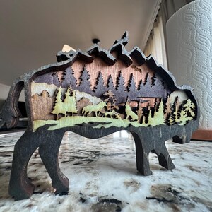 Handcrafted wooden bison wall art featuring a layered forest and mountain scene with wildlife details. Measures 6"x6"x3/8", perfect for rustic, farmhouse, or cabin décor. A unique gift for nature lovers and wildlife enthusiasts.