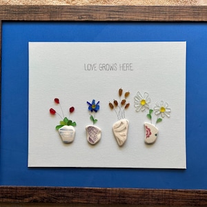 May include: A framed wall art piece featuring the text LOVE GROWS HERE above four small vases made of ceramic shards. Each vase holds floral arrangements crafted from sea glass, wire, and small stones in red, blue, white, and yellow colors.