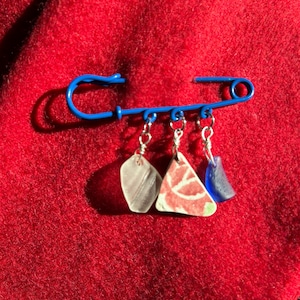 May include: A blue safety pin brooch with three dangling charms. The charms include a clear, a red and white patterned, and a blue piece. The brooch is set against a red, textured background.