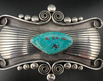 May include: A silver belt buckle with a turquoise stone set in the center. The buckle is decorated with intricate swirls and bead accents.
