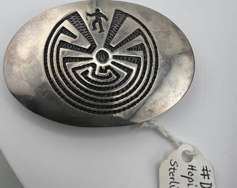 May include: An oval sterling silver belt buckle featuring an etched Hopi maze design with a central figure. A white paper tag attached with string displays the handwritten text: # DA 10 Hopi Buckle Sterling Silver.