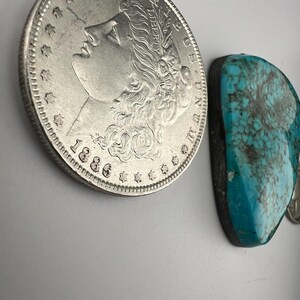 May include: A silver Morgan dollar coin from 1886 next to a turquoise cabochon. The coin features the profile of Lady Liberty with the inscription "E PLURIBUS UNUM" and "LIBERTY".
