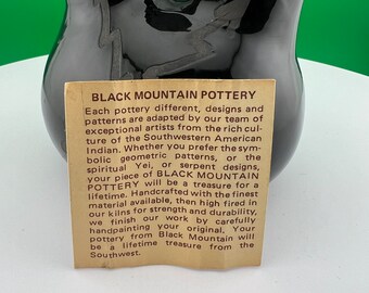 May include: A glossy black pottery vase with a unique, curved design. The vase features a lightning bolt design and a paper tag with the text "BLACK MOUNTAIN POTTERY". The pottery is handcrafted and from the Southwest.