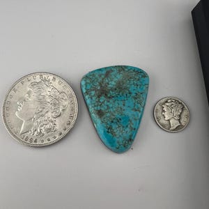 May include: A turquoise cabochon with a triangular shape and a mottled pattern. The cabochon is surrounded by two silver coins, one with a portrait of a woman and the other with a portrait of a man.