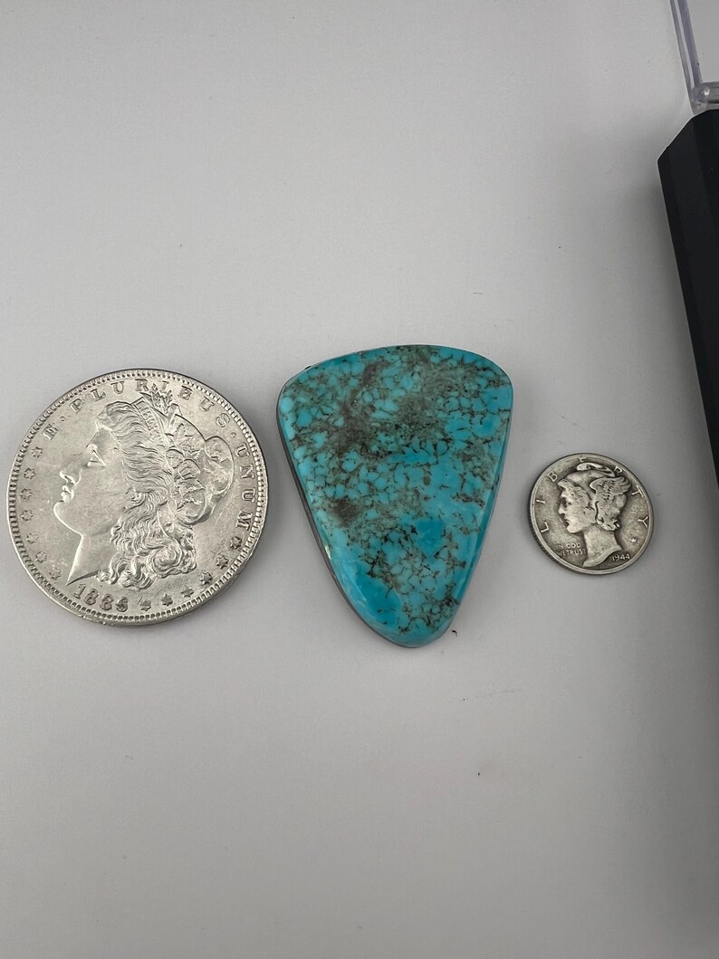 May include: A turquoise cabochon with a triangular shape and a mottled pattern. The cabochon is surrounded by two silver coins, one with a portrait of a woman and the other with a portrait of a man.