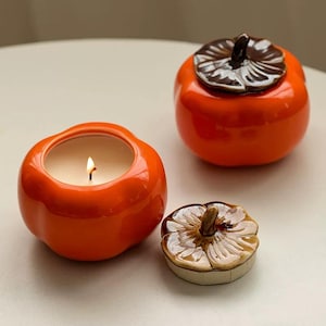 May include: Orange ceramic candle holder shaped like a persimmon fruit, with a lit candle inside. The lid is off, revealing the candle. Another persimmon-shaped container with a lid is in the background. The lid is brown and tan.