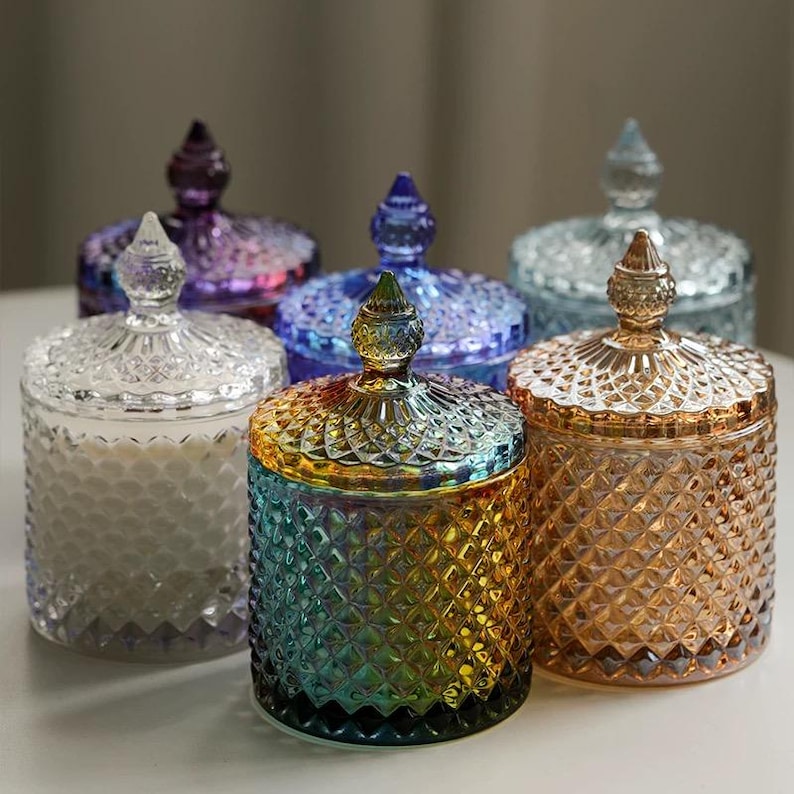 May include: Six decorative glass jars with lids. The jars are in various colors, including clear, purple, blue, and gold. Each jar has a diamond pattern and a decorative finial on the lid. The jars are arranged on a white surface.