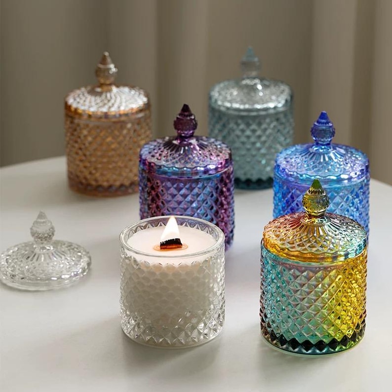 May include: A collection of decorative glass candle jars in various colors, including gold, purple, blue, and teal. Each jar has a diamond pattern and a matching lid with a decorative knob. One jar is lit, showing a burning wick.