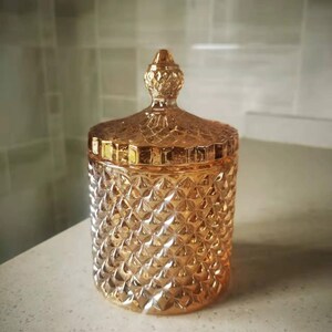 May include: A decorative, amber-colored glass jar with a diamond pattern and a matching lid. The jar has a cylindrical shape and a textured surface. The lid is topped with a small, ornate finial. The jar is likely used for storage or display.