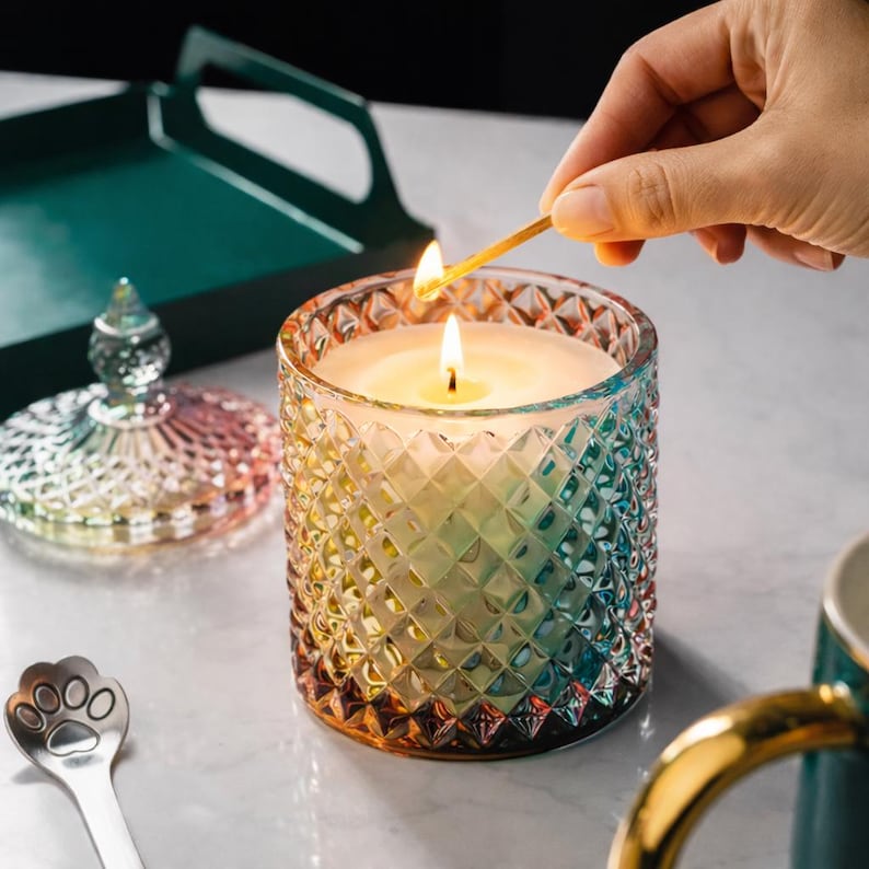 May include: A lit, multi-colored glass candle with a diamond pattern. The candle is being lit with a match. A matching lid and a paw-shaped spoon are nearby. The candle's colors transition from orange to blue.