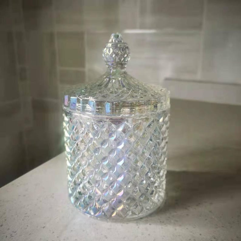 May include: A clear, iridescent glass jar with a diamond-patterned exterior and a decorative lid. The jar is cylindrical with a rounded base and a matching lid with a knob. The glass reflects light, creating a rainbow effect.