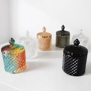 May include: Six decorative glass jars with lids. The jars are in various colors, including clear, gold, black, and a rainbow gradient. Each jar has a diamond pattern and a decorative knob on the lid. The jars are arranged on a white surface.