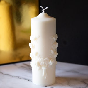 May include: A white pillar candle with a floral design. The candle is decorated with raised floral details and has a white wick.