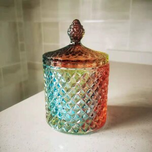 May include: A decorative glass jar with a lid. The jar is a vibrant mix of colors, including blue, green, orange, and red. The jar has a diamond pattern and a decorative knob on the lid. The jar is likely used for storage or decoration.