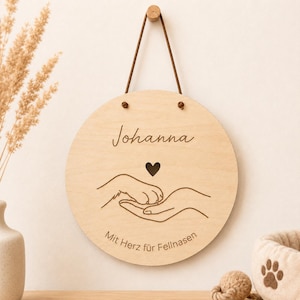 Könnte beinhalten: Ein rundes Wandschild aus hellem Holz mit der Gravur des Namens Johanna, einem kleinen Herz und einer Strichzeichnung einer menschlichen Hand, die eine Hundepfote hält. Der Text Mit Herz für Fellnasen ist unten eingraviert. Es hängt an einer braunen Kordel.