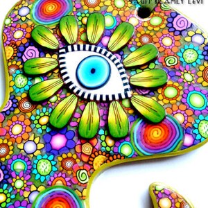 May include: A colorful, handcrafted decorative piece with a floral design and an eye-shaped centerpiece. The piece features a vibrant mix of colors, including green, blue, yellow, orange, and purple. The text "SIGALIOT SAGIT BRAMLY LEVI" is visible.
