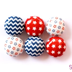 May include: Six round wooden ornaments with red, white, and blue patterns. Three ornaments have a white background with red stars and a blue outline. Three ornaments have a blue chevron pattern on a white background.