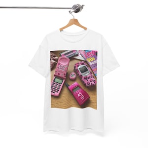 May include: White t-shirt featuring a collection of pink flip phones, a leopard print phone, a cassette tape, and a magazine with the text "90s Badd". The shirt has a vintage, retro aesthetic.