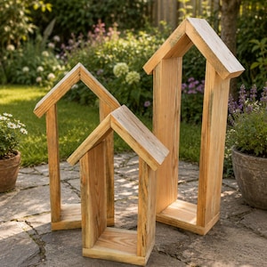 May include: Three house-shaped wooden frames of varying heights, constructed from light-colored natural wood with visible grain patterns. Each frame features a peaked roof design and an open center, standing upright on a stone patio surface.