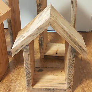 May include: A small, open-fronted structure crafted from light-colored, unfinished wood. It features a peaked, gabled roof with two angled panels meeting at a central ridge, supported by two vertical side walls and a flat, rectangular base.