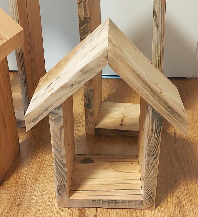 May include: A small, open-fronted structure crafted from light-colored, unfinished wood. It features a peaked, gabled roof with two angled panels meeting at a central ridge, supported by two vertical side walls and a flat, rectangular base.