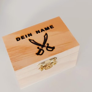 Könnte beinhalten: Eine Holzkiste mit einem Scharnierdeckel und einem Verschluss. Die Kiste ist mit dem Text "DEIN NAME" und einem Paar gekreuzter Schwerter graviert.