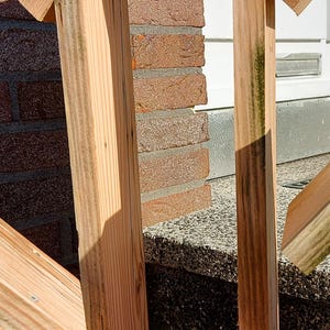 May include: A tall, rectangular wooden frame with a peaked, gabled roof structure. The object is constructed from light-colored, unfinished pine wood planks with visible grain patterns and some green-tinted weathering along the edges.