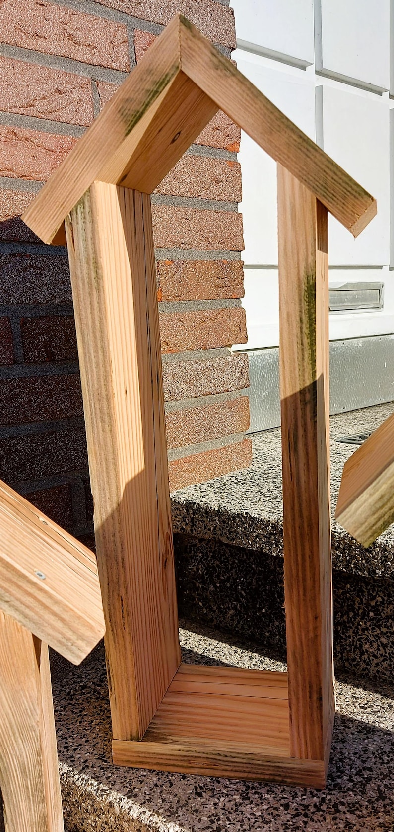 May include: A tall, rectangular wooden frame with a peaked, gabled roof structure. The object is constructed from light-colored, unfinished pine wood planks with visible grain patterns and some green-tinted weathering along the edges.
