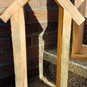 May include: A tall, narrow, house-shaped wooden frame constructed from light-colored, unfinished planks. The structure features two vertical side panels, a flat base, and a peaked roof with two angled boards meeting at a central point at the top.