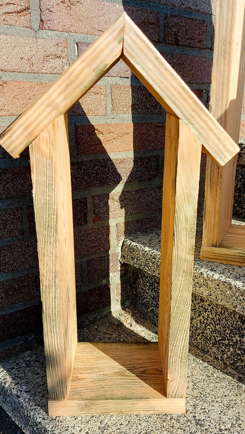 May include: A tall, narrow, house-shaped wooden frame constructed from light-colored, unfinished planks. The structure features two vertical side panels, a flat base, and a peaked roof with two angled boards meeting at a central point at the top.