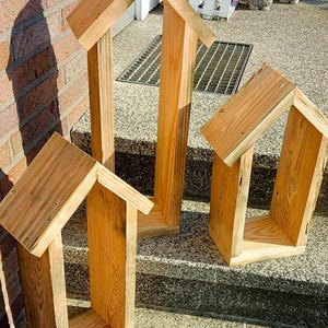 May include: Three house-shaped wooden frames constructed from light-colored natural wood planks. Each frame features a peaked roof design with vertical side walls and a flat base, showcasing visible wood grain patterns and metal screw fasteners.