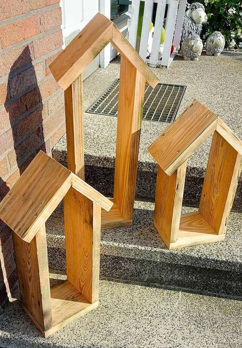 May include: Three house-shaped wooden frames constructed from light-colored natural wood planks. Each frame features a peaked roof design with vertical side walls and a flat base, showcasing visible wood grain patterns and metal screw fasteners.