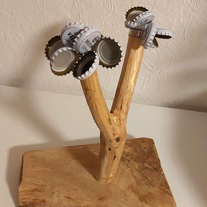 Könnte beinhalten: Ein Flaschenöffner aus Holz in Baumform mit natürlicher Holzoptik. Der Flaschenöffner hat eine Basis aus einem dicken Stück Holz und zwei Äste, die sich nach oben erstrecken. Die Äste sind mit Flaschenverschlüssen verziert.