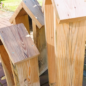 May include: Three tall, rectangular wooden structures with pitched roofs, constructed from light-colored natural wood planks. Each piece features visible wood grain, vertical side panels, and small metal screws securing the roof sections together.