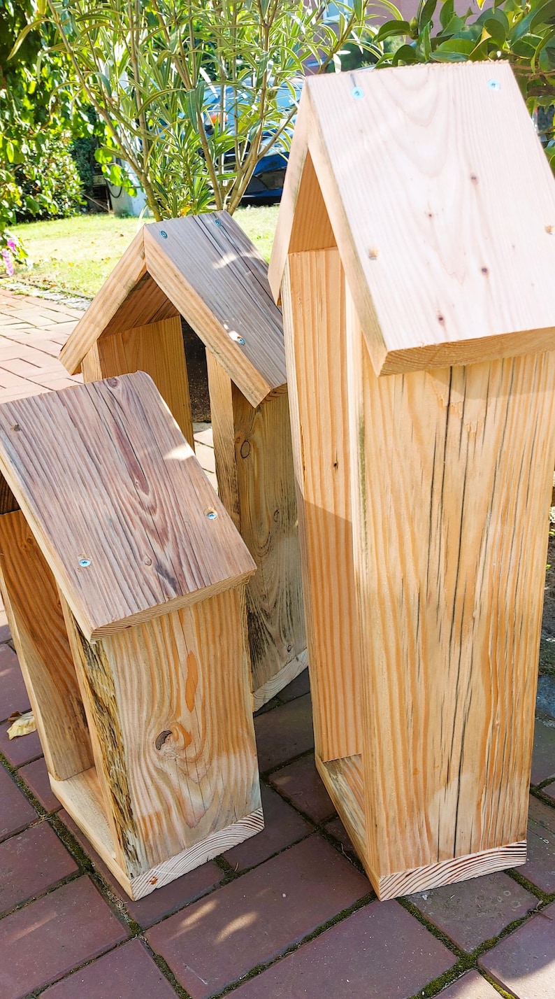 May include: Three tall, rectangular wooden structures with pitched roofs, constructed from light-colored natural wood planks. Each piece features visible wood grain, vertical side panels, and small metal screws securing the roof sections together.